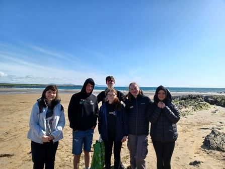 A group of six teenagers, volunteering with North Wales Wildlife Trust, stood on a beach, with the sea visible behind them. Anna is stood in the middle of the group