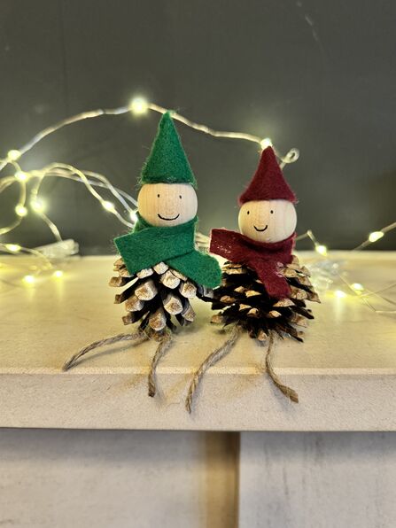Two elves made out of pinecones set on a shelf, with fairy lights behind them