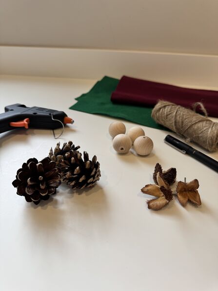 The collection of materials needed to make pinecone elves: a glue gun, pine cones, felt, twine, wooden beads, a fineliner and seed pods